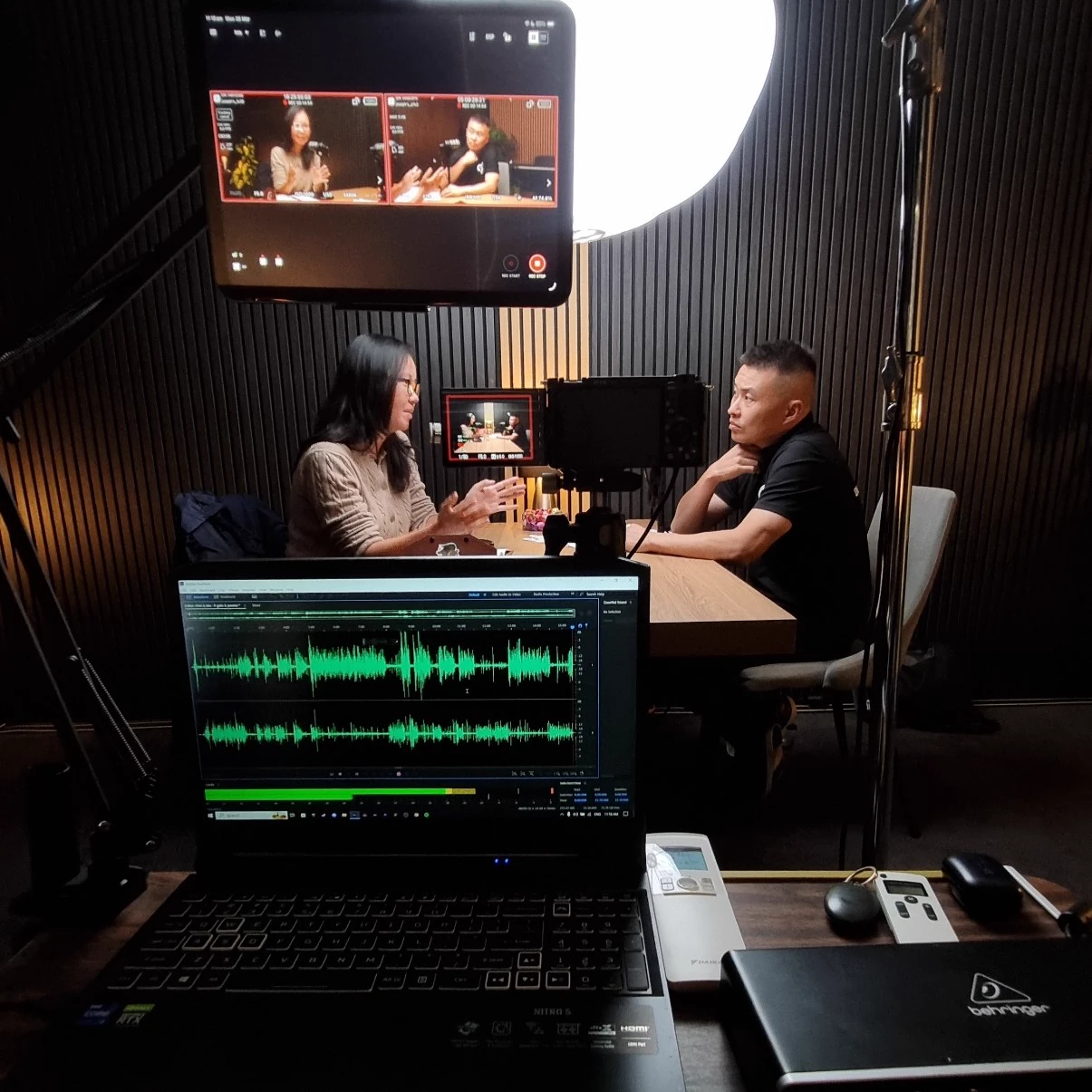 Man and woman recording an interview at a table with waveforms and a camera visible